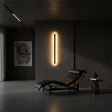 A modern, minimalist room features a black leather chaise lounge, a small round side table with a vase, abstract wall art, and the Averra LED Extra Large Round Alabaster Wall Light in soft white & brass glowing on a dark gray wall.