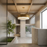 A modern interior with high ceilings, floor-to-ceiling windows, light wood paneling, a built-in planter featuring a small tree, minimalist white and grey walls, and the Royal 6-12 Crystal Light Chandelier - Gold as a striking centerpiece.
