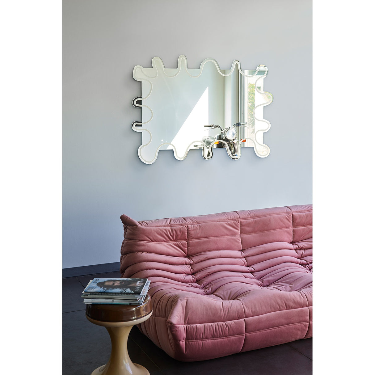 Wiggle Scalloped Edge Wall Mirror 133cm with a wavy, frameless glass outline and engraved inner border, hung above a pink sofa in a living room setting.