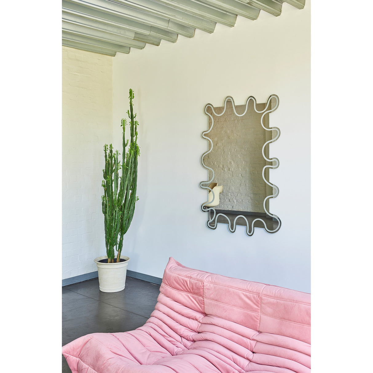Wiggle Scalloped Edge Wall Mirror 133cm with a wavy, frameless glass outline and engraved inner border, mounted on a white wall above a pink sofa in a living room setting.
