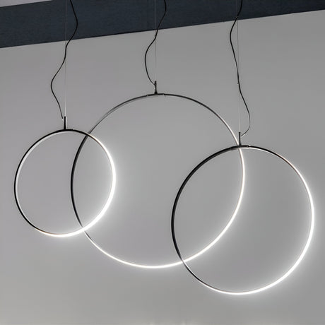 The Claria LED Suspension Light 29W 3000K - Black, 44cm showcases a minimalist design with three interlocking circular rings of different sizes, elegantly suspended from the ceiling by black cords. This fixture emits a bright and even glow, highlighting its modern design against a plain gray background and creating an elegant ambiance.