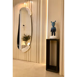 Calco Sculpted Full Length Wall Mirror 166cm in White, an oval mirror with irregular white moulded forms at top and bottom, shown in a hallway beside a dark pedestal with sculpture.