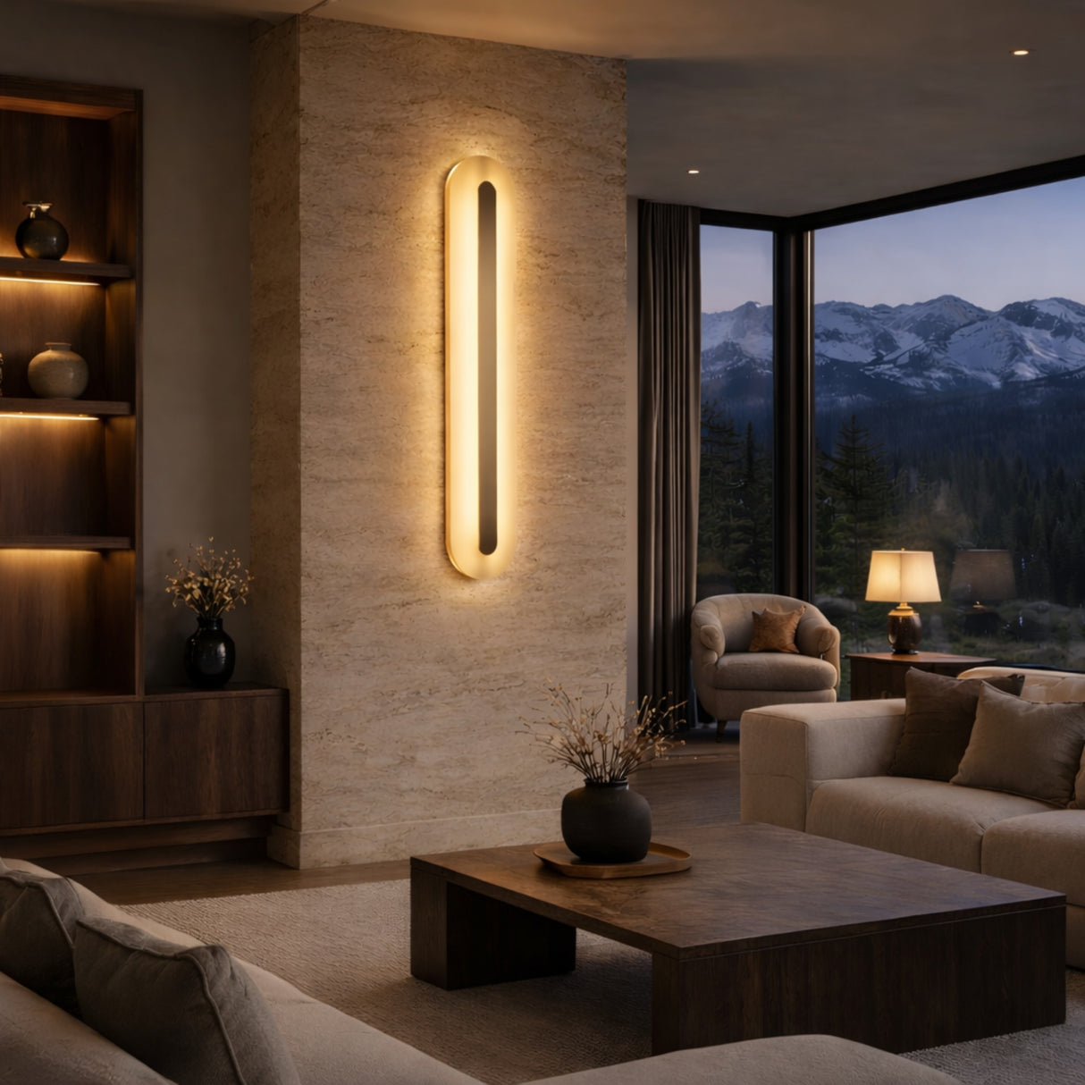 A modern living room with beige sofas, a wooden coffee table, and built-in shelves. The Averra LED Extra Large Rectangle Alabaster Wall Light in soft white and brass glows on a stone accent wall. Large windows reveal mountains at dusk.