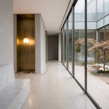 A modern hallway with large floor-to-ceiling glass windows overlooks a tree-lined courtyard with beige stone floors, a marble bench, and the Vetra 1 Light Opal Glass Floor Lamp - Gold in a brass alcove.