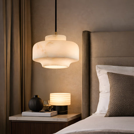 A cozy bedroom features the Doreth 1 Light Large Dome Alabaster Pendant in soft white and brushed brass above a wooden nightstand, paired with a small table lamp beside a bed with neutral bedding and pillows.