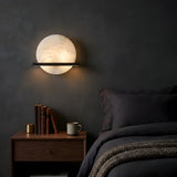 A modern bedroom with dark walls features the Caldris LED Circular Alabaster Wall Light in Matte Black & Soft White above a wooden nightstand, books, a brass object, mug, and a neatly made bed with dark bedding and a knitted blanket.