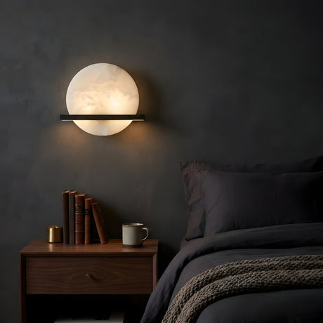 A modern bedroom with dark walls features the Caldris LED Circular Alabaster Wall Light in Matte Black & Soft White above a wooden nightstand, books, a brass object, mug, and a neatly made bed with dark bedding and a knitted blanket.