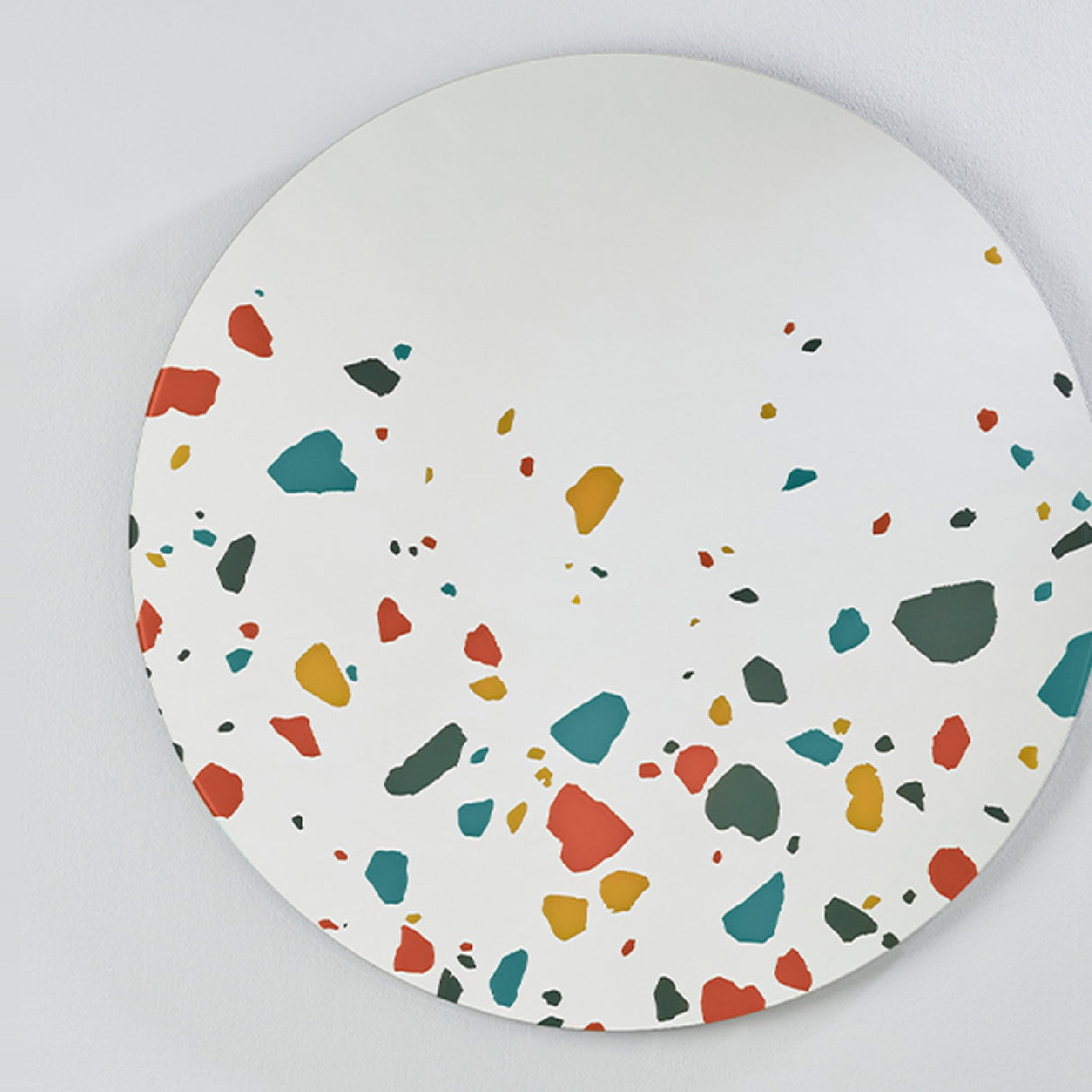 Terrazzo Round Printed Glass Wall Mirror 100cm, a frameless round mirror with red, teal, ochre and grey terrazzo chip print clustered at the lower edge, mounted on a light grey wall.