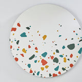 Terrazzo Round Printed Glass Wall Mirror 100cm, a frameless round mirror with red, teal, ochre and grey terrazzo chip print clustered at the lower edge, mounted on a light grey wall.