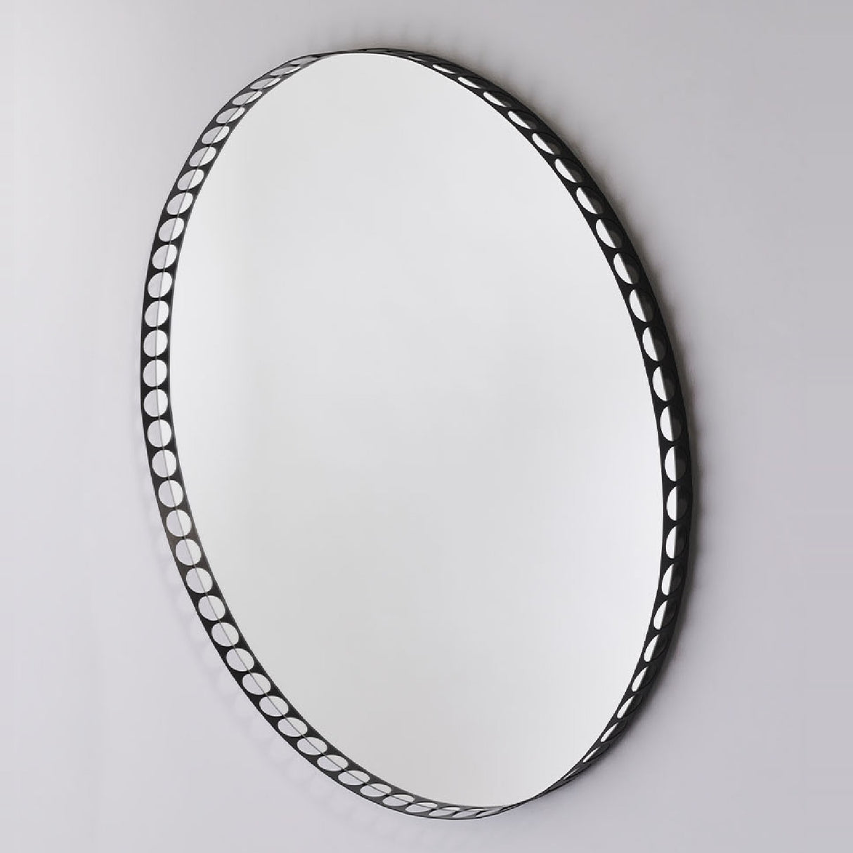 Round wall mirror with a black metal rim perforated by a row of round cut-out holes, shown at an angle against a plain light grey background.