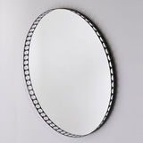 Round wall mirror with a black metal rim perforated by a row of round cut-out holes, shown at an angle against a plain light grey background.