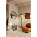 Round wall mirror with a black perforated metal rim, reflecting a green dome floor lamp, in a living room with a rug, side table, and armchair.