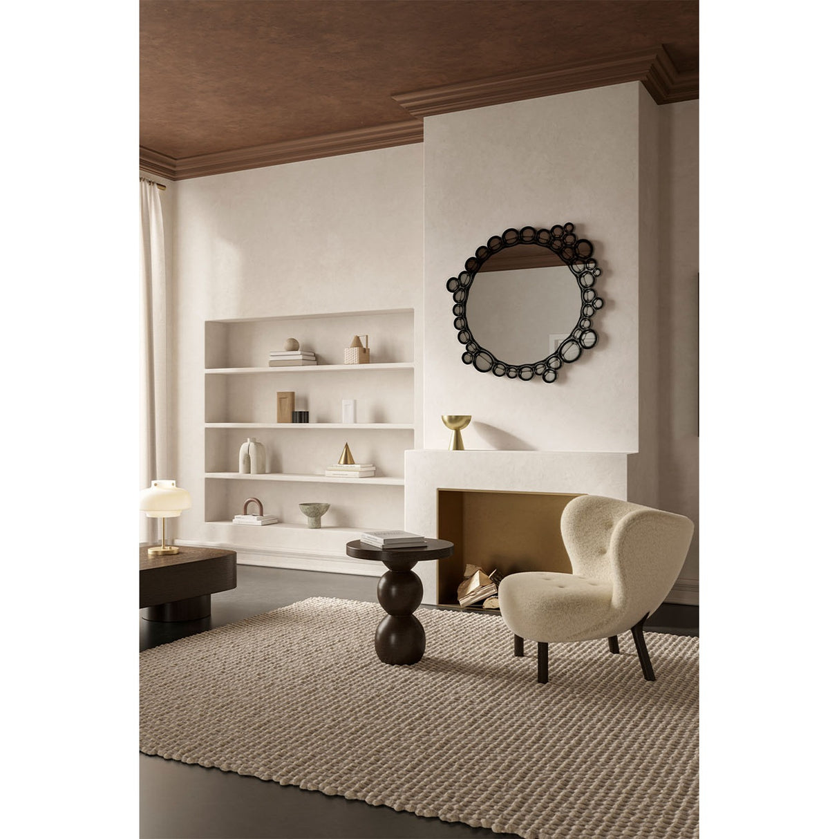 Round wall mirror with a black frameless border of overlapping printed glass bubbles, mounted above a fireplace in a living room with a shelving unit and cream armchair nearby.