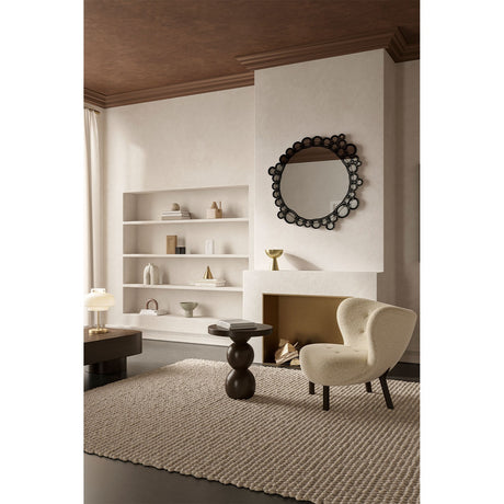 Round wall mirror with a black frameless border of overlapping printed glass bubbles, mounted above a fireplace in a living room with a shelving unit and cream armchair nearby.