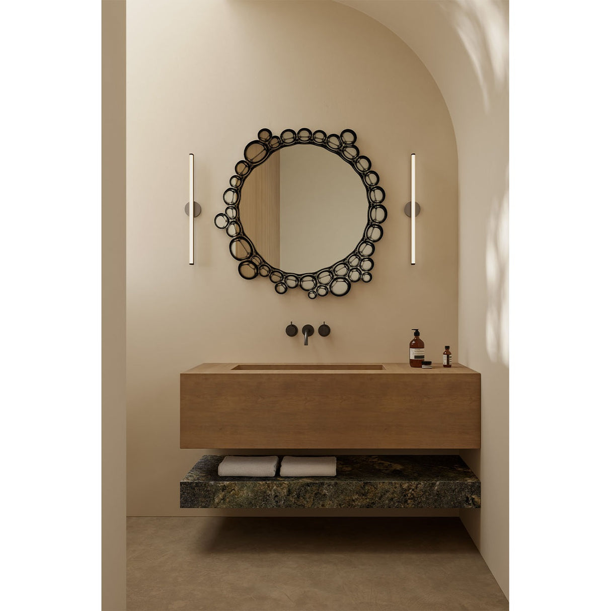 Round wall mirror with a black bubble-patterned glass frame of overlapping circles, hung above a wood vanity with wall-mounted taps in a bathroom setting.