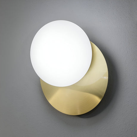 The Halion Wall Light - Brass is a modern lamp featuring overlapping circular shapes, with a white shade mounted on a larger brass disc against a grey wall, effortlessly blending minimalist and elegant design.