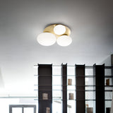 A modern interior features an elegant Halion 3 Light Flush in brass, adorned with globe-shaped bulbs and metal accents. Below it, an open shelf with glass dividers showcases various items, while a window in the background invites natural light to illuminate the room.