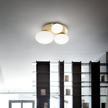 A modern interior features an elegant Halion 3 Light Flush in brass, adorned with globe-shaped bulbs and metal accents. Below it, an open shelf with glass dividers showcases various items, while a window in the background invites natural light to illuminate the room.
