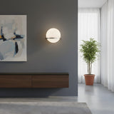 A modern minimalist living room with the Caldris LED Circular Alabaster Wall Light in satin nickel & soft white, an abstract painting, floating wooden cabinet, potted plant, white curtains, and streaming natural sunlight.