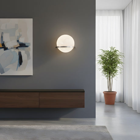 A modern minimalist living room with the Caldris LED Circular Alabaster Wall Light in satin nickel & soft white, an abstract painting, floating wooden cabinet, potted plant, white curtains, and streaming natural sunlight.