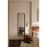 Calligra Curved Metal Wall Mirror 168cm in black, oval-shaped with a curved frame, leaning against a bedroom wall beside a wooden side table and bed.