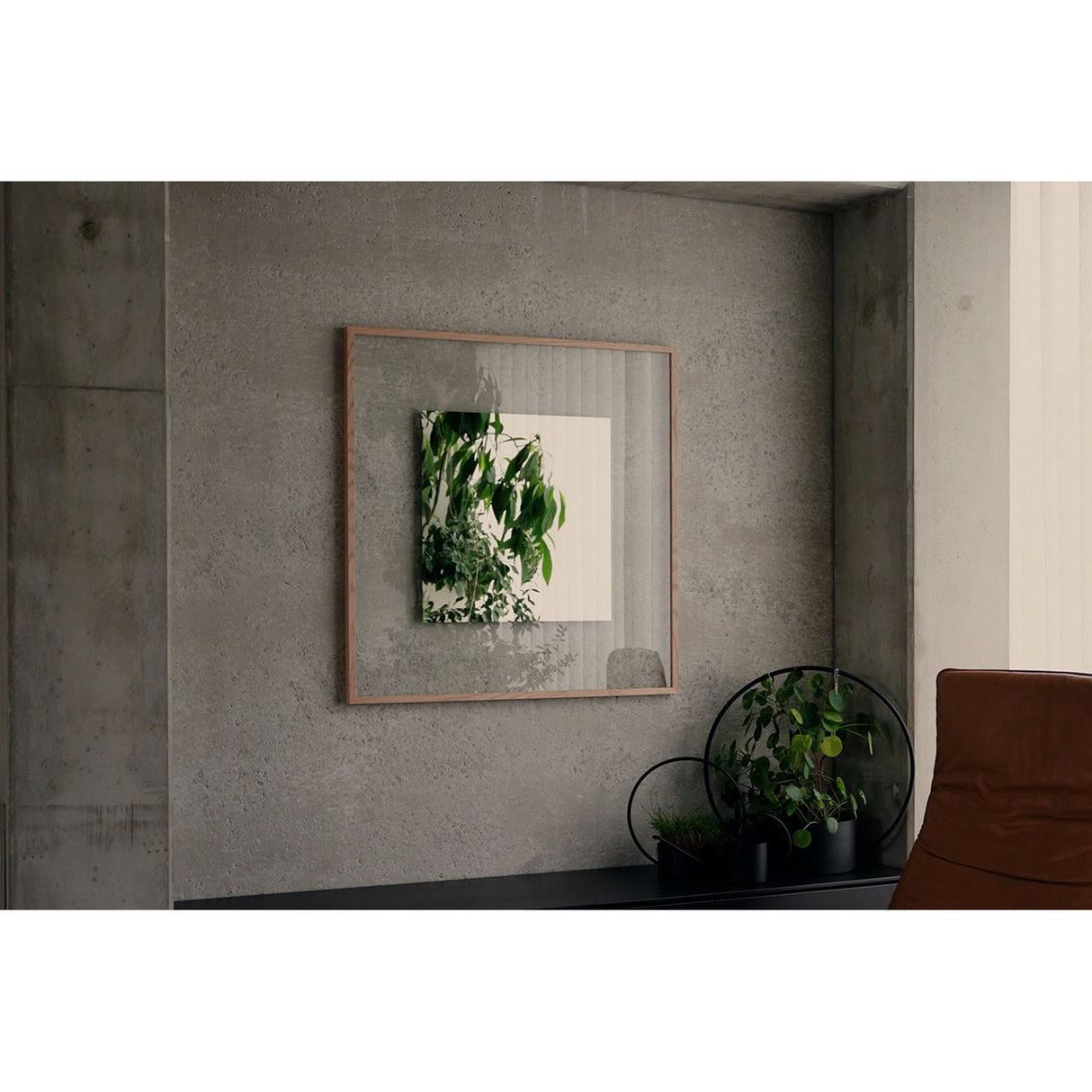 Passepartout Square wall mirror in solid oak frame, 105cm, mounted on a concrete wall, reflecting green foliage, with a sideboard and potted plants below.