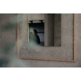 Passepartout Square wall mirror in solid oak, mounted on a grey concrete wall, reflecting a blurred figure and interior with a soft-edged, fading mirrored centre.