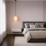 Minimalist bedroom with a neatly made bed featuring beige and blue pillows, a soft beige comforter, the Bellorin 1 Light Large Alabaster Single Pendant Light in Soft White & Brushed Brass, sheer white curtains, and dark wood flooring.