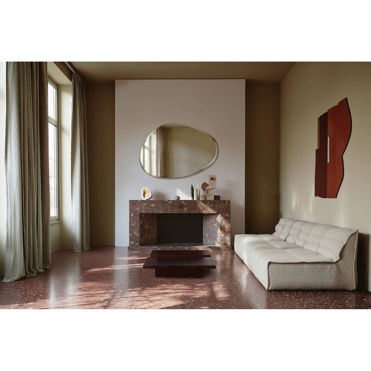 Frameless organic-shaped wall mirror with bevelled edge, 136cm, mounted above a marble fireplace in a living room with a cream sofa and terrazzo floor.