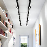 A modern hallway with white walls and a light wooden floor is decorated with bookshelves filled with books on the left and a framed picture on the right wall. The ceiling is enhanced by integrated LED technology featuring the Helionis LED Track Square Spotlight 22W 3000K in black, while a window at the end provides a view of greenery outside.