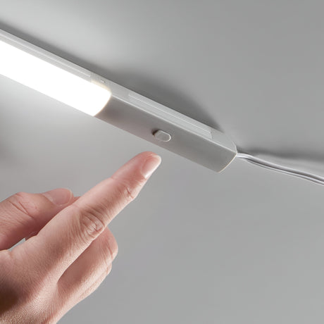 A person's hand is poised to press the Nocturne Button Switch on a sleek, modern under-cabinet LED lighting system. The light glows softly, connected with a clearly visible power cord.