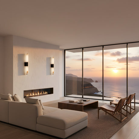 A modern living room with a white sectional, wooden chairs, and a minimalist fireplace showcases the Fiora LED Alabaster Wall Light – Matte Black & Soft White. Floor-to-ceiling windows offer sunset ocean views as warm light fills the serene space.