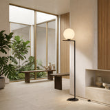 A modern, minimalist living space features large windows, indoor plants, wooden benches, and the Vetra 1 Light Opal Glass Floor Lamp - Black. Light wood and neutral tones create a calm atmosphere enhanced by contemporary lighting.