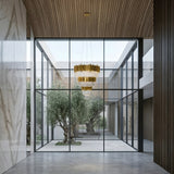 Niori Royal 30-Light Tiered Crystal Chandelier in gold hanging from a wood-slat ceiling above a glass-walled courtyard with olive trees and marble walls.