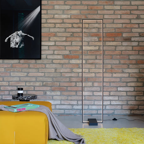 A modern living space with a brick wall showcases the Prios LED Floor Lamp 30W 3000K in black, accompanied by a black and white framed photo. The minimalist design elements include yellow cushioned seating draped with a gray blanket and a small round table decorated with books and a candle.