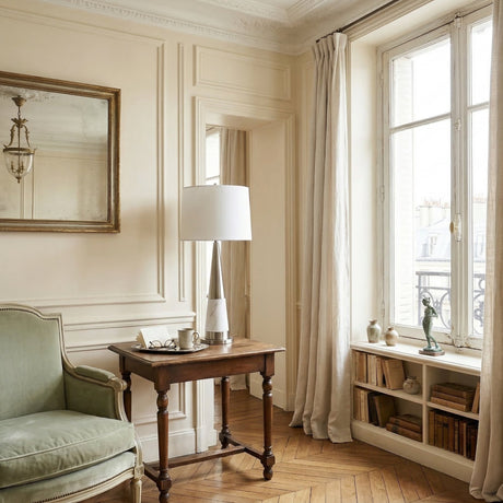 Elegant Parisian sitting room with cream wall paneling, herringbone parquet floors, sage green velvet armchair, antique wooden side table topped with the Velour 1 Light Cone Marble Table Lamp in white & brass, gilt mirror, and tall windows with linen drapes.