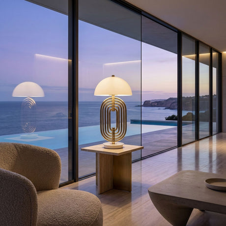 A modern living room with large glass windows overlooks the ocean at sunset. The Ascot 4 Light Large Table Lamp in white and gold sits on a side table by a textured armchair, its warm glow enhancing the peaceful atmosphere.