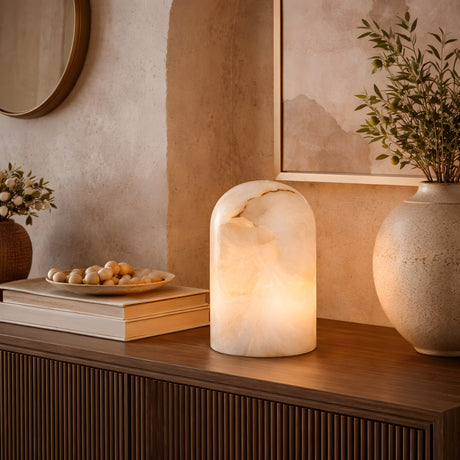The Virelle LED Arched Dome Alabaster Table Lamp in Soft White glows on a wooden sideboard beside stacked books, beaded plate, and a leafy vase, set against a beige textured wall with a round mirror and framed art.