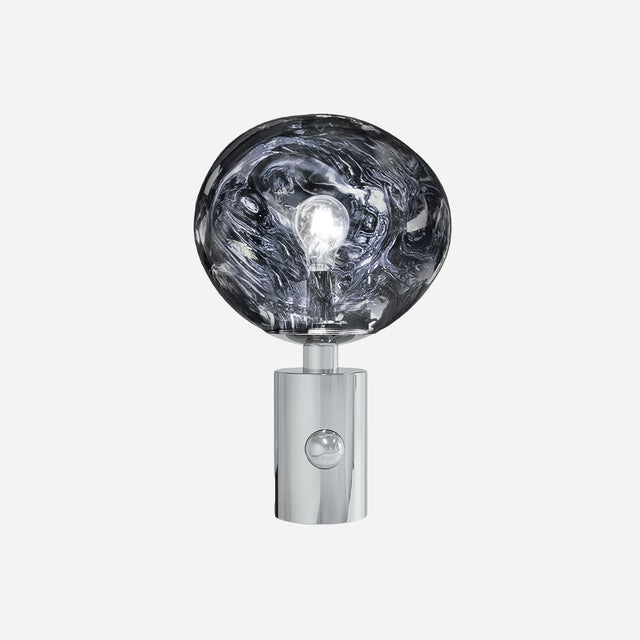 The Ex-Display Lava Table Lamp - Silver is a contemporary lighting piece with a shiny, cylindrical chrome base and a large, uniquely shaped clear glass bulb that showcases the lightbulb within—all set against a plain white background.