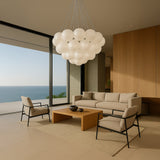 A modern living room with ocean views, neutral furniture, a wooden coffee table, and the Orb 17 Light Cluster Suspension - Silver featuring frosted white glass spheres hanging from the ceiling.