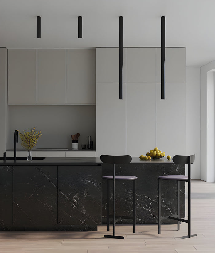 Modern kitchen with sleek, minimalist design. Features a black marble island with high stools, a white cabinet backdrop, and elegant black pendant lights. Yellow lemons are arranged on the countertop, adding a pop of color.