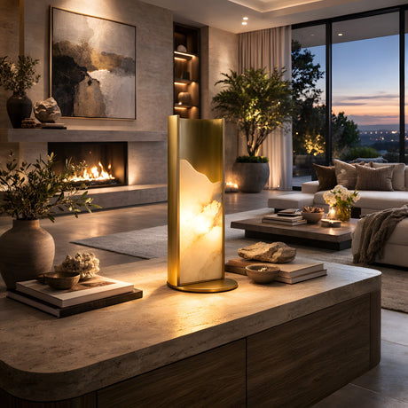 Niori Caelith LED carved alabaster table lamp in brushed brass glowing on a stone coffee table in a modern living room with fireplace and sunset view.
