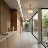 A modern hallway with wooden paneling features the Brinex 1 Light Alabaster Single Pendant Light in Soft White & Brushed Brass, its glow complementing large glass windows, a marble countertop with a vase and books, and views of a bamboo garden outside.