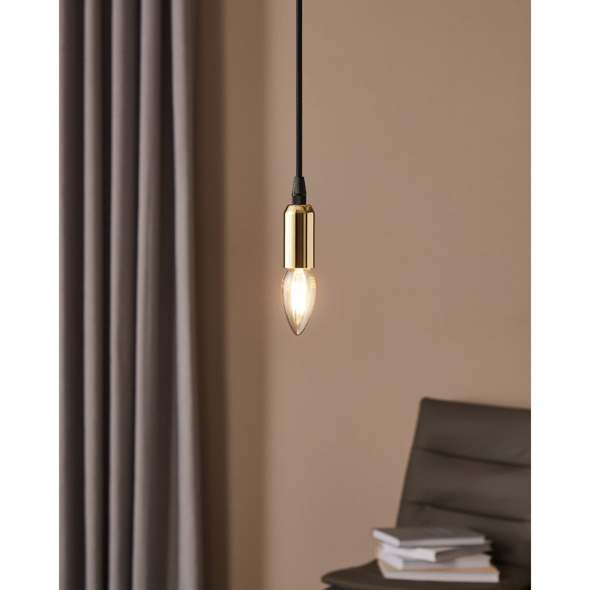 Pendant light with brass fitting holding a lit E14 LED candle bulb, clear glass with visible filaments, shown against a curtain in a living room setting.