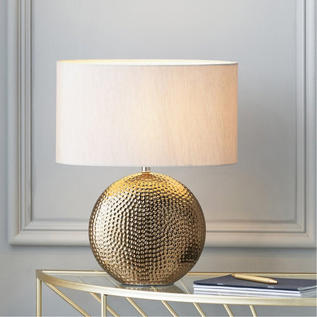 Mabel Bronze Dot Textured Ceramic Table Lamp with an oval cream fabric shade, shown on a glass console table in a living room setting.