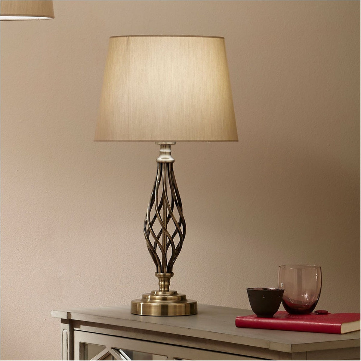 Jenna Antique Brass Gold Twist Detail Table Lamp with a spiral metal base and tapered fabric shade, styled on a sideboard beside a red book and glassware.