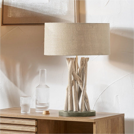 Derna Driftwood Table Lamp with a natural jute drum shade and twisted driftwood base, shown on a wooden sideboard beside glassware in a bright living room setting.