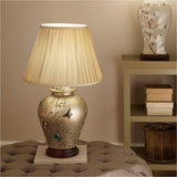Papillon Butterfly Ceramic Table Lamp, 61cm, with gold ceramic base featuring a floral and butterfly design, pleated cream fabric shade, shown on a footstool beside a shelving unit.