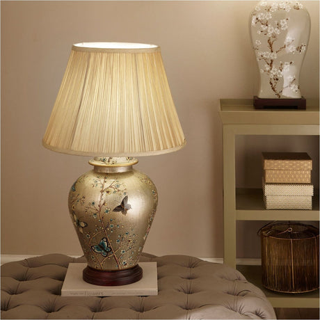 Papillon Butterfly Ceramic Table Lamp, 61cm, with gold ceramic base featuring a floral and butterfly design, pleated cream fabric shade, shown on a footstool beside a shelving unit.