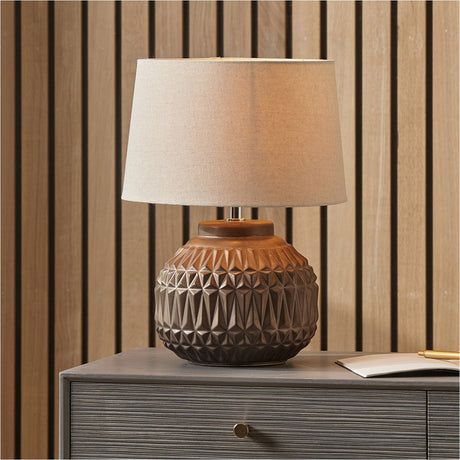 Anneli table lamp with bronze ceramic base in geometric Aztec texture and cream linen tapered shade, shown on a grey dresser against wood slat panelling.